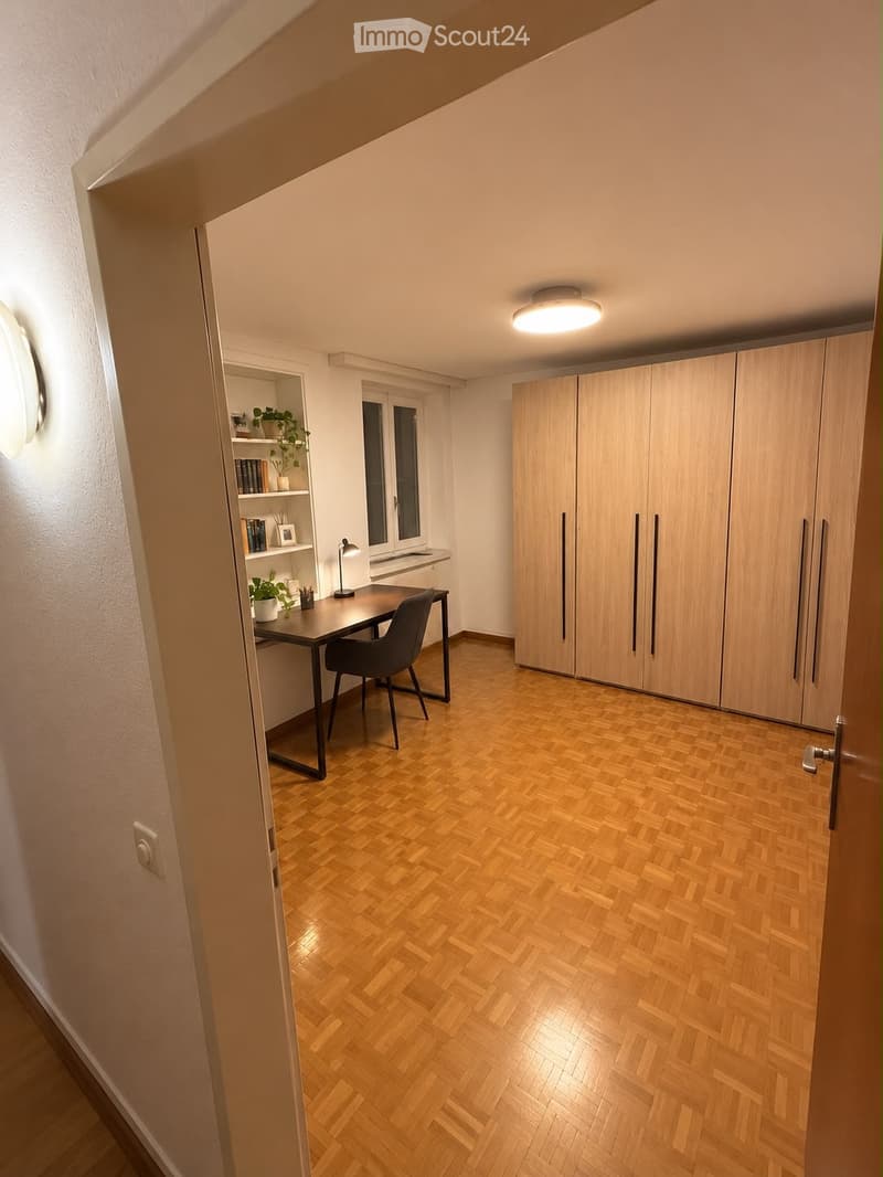 Modern, well-lit room with wooden herringbone flooring, large built-in wardrobe, desk with chair, bookshelf, and small plant.