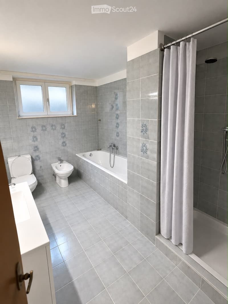 Modern bathroom with grey tiles, a bathtub, toilet, bidet, and a shower area with a curtain.