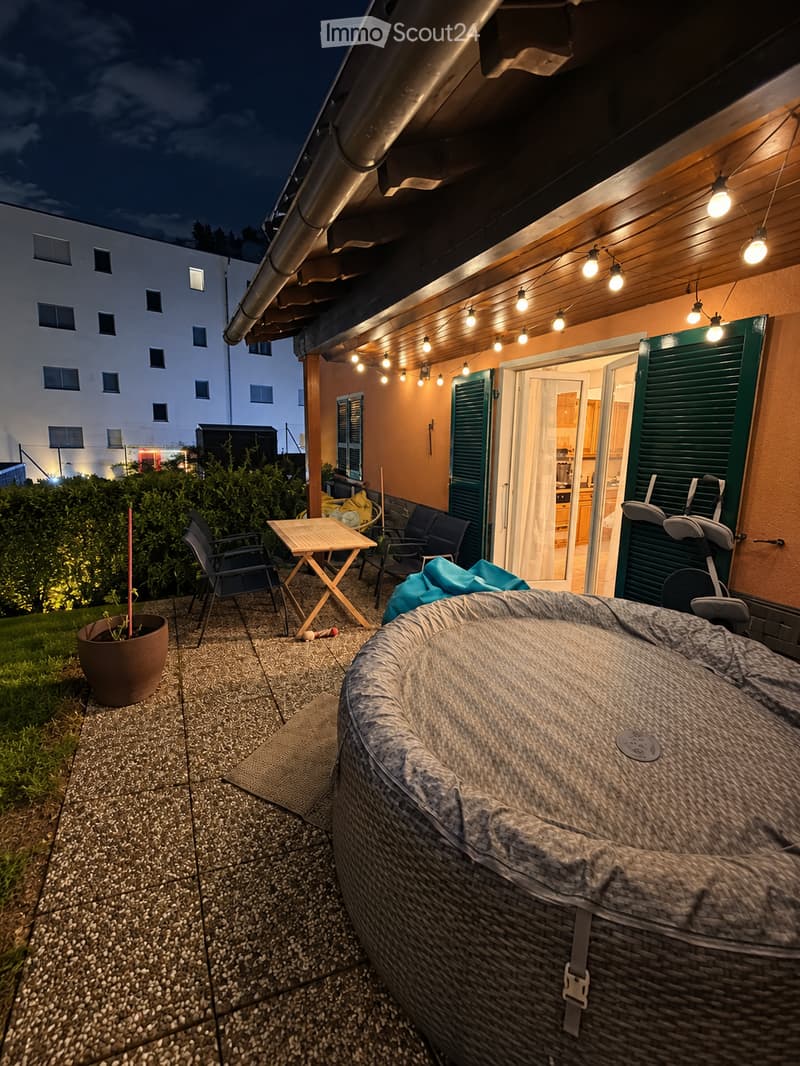 Outdoor patio area with a hot tub, small table, chairs, and a potted plant, illuminated by string lights.