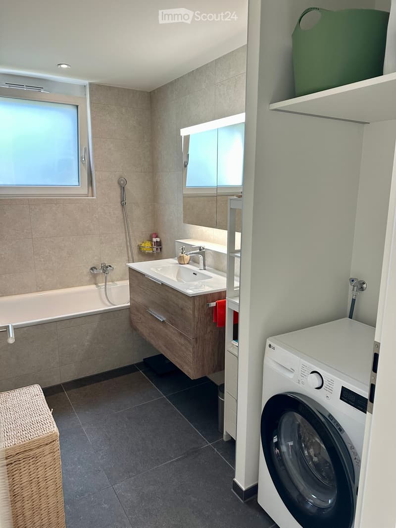 Modern bathroom with a bathtub, sink, mirror, and a private washing machine. Features include a wicker basket, towels, and a sleek design with wooden cabinetry.