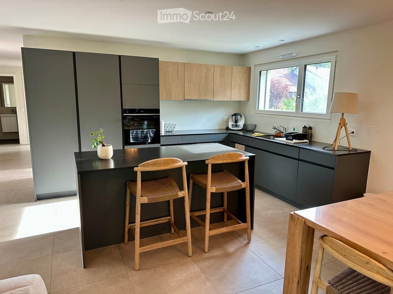 Modern kitchen with dark grey cabinets, light wooden upper cabinets, island with black countertop, two wooden stools, small table with chairs, and a potted plant on the island.
