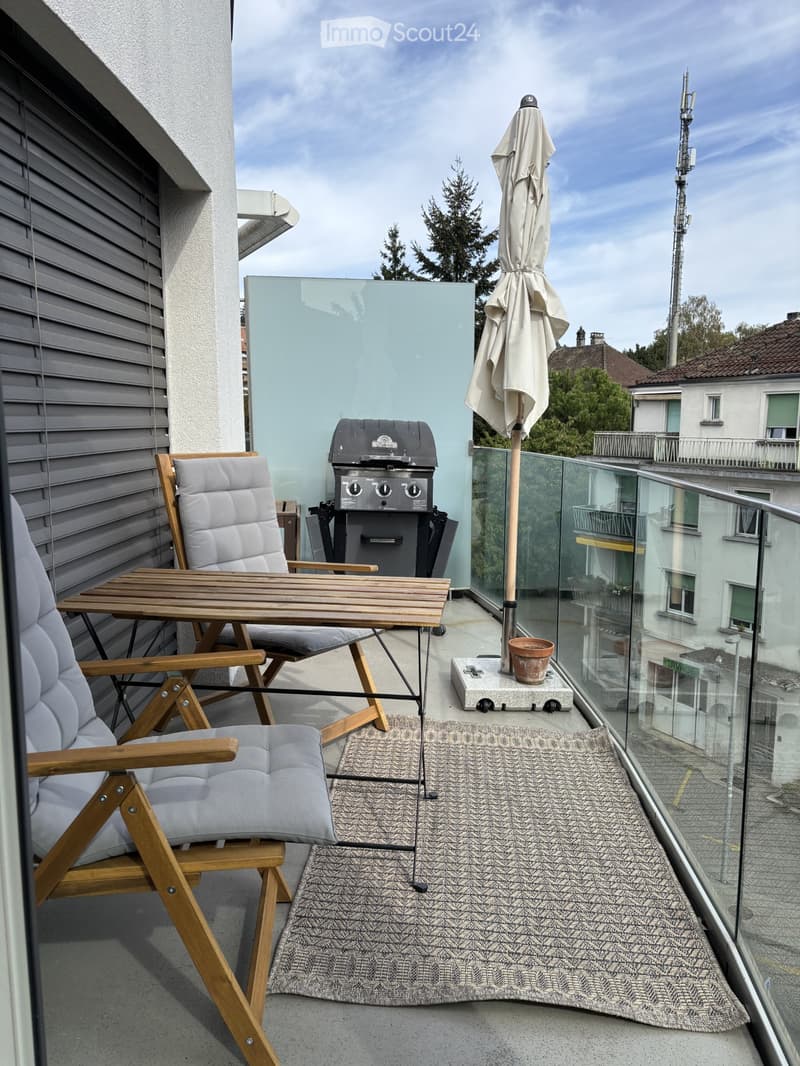 Balcony with a wooden table, two chairs, a grill, an umbrella, and a rug.