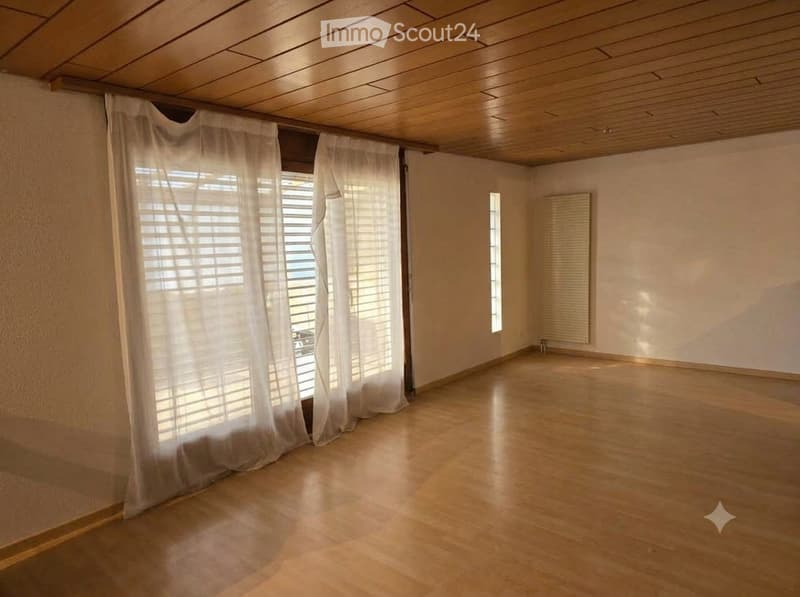 Empty room with wooden flooring, white walls, large window with blinds, and a radiator on one wall.
