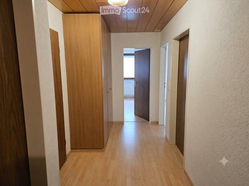 A clean, well-lit hallway with wooden flooring, white walls, and a wooden cabinet on the left side. The hallway leads to an open doorway at the end.