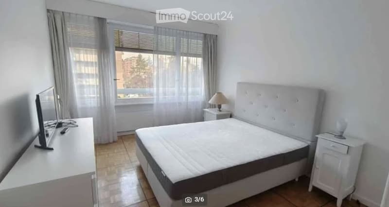 Modern bedroom with a large bed, nightstands, a lamp, and a TV screen on a stand. The room has large windows with curtains and a tiled floor.