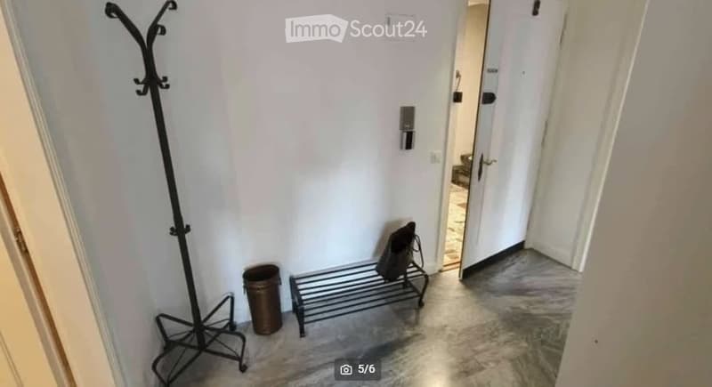 A narrow hallway with white walls, a coat rack, a small bench with a bag, and a trash bin. The floor is tiled, and there is an open doorway leading to another room.