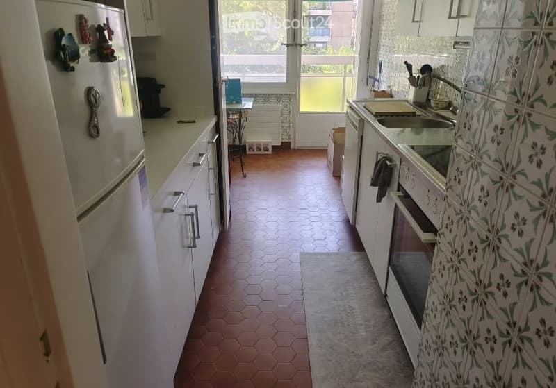 A compact kitchen with white cabinets, a refrigerator, a sink, and an oven. The floor is tiled in a hexagonal pattern, and there is a small table and stool near the window.