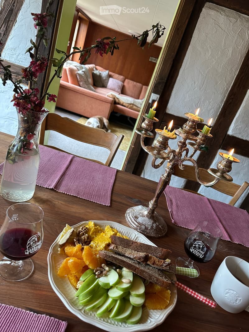 Breakfast table set with a plate of fruit, bread, and cheese, accompanied by wine glasses, a candle holder, and a vase with flowers. A pink sofa is visible in the background.