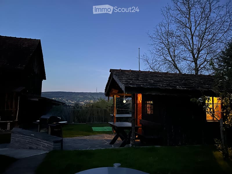 A serene outdoor area with a wooden cabin, a picnic table, a bench, and a barbecue, set against a backdrop of a scenic valley view.