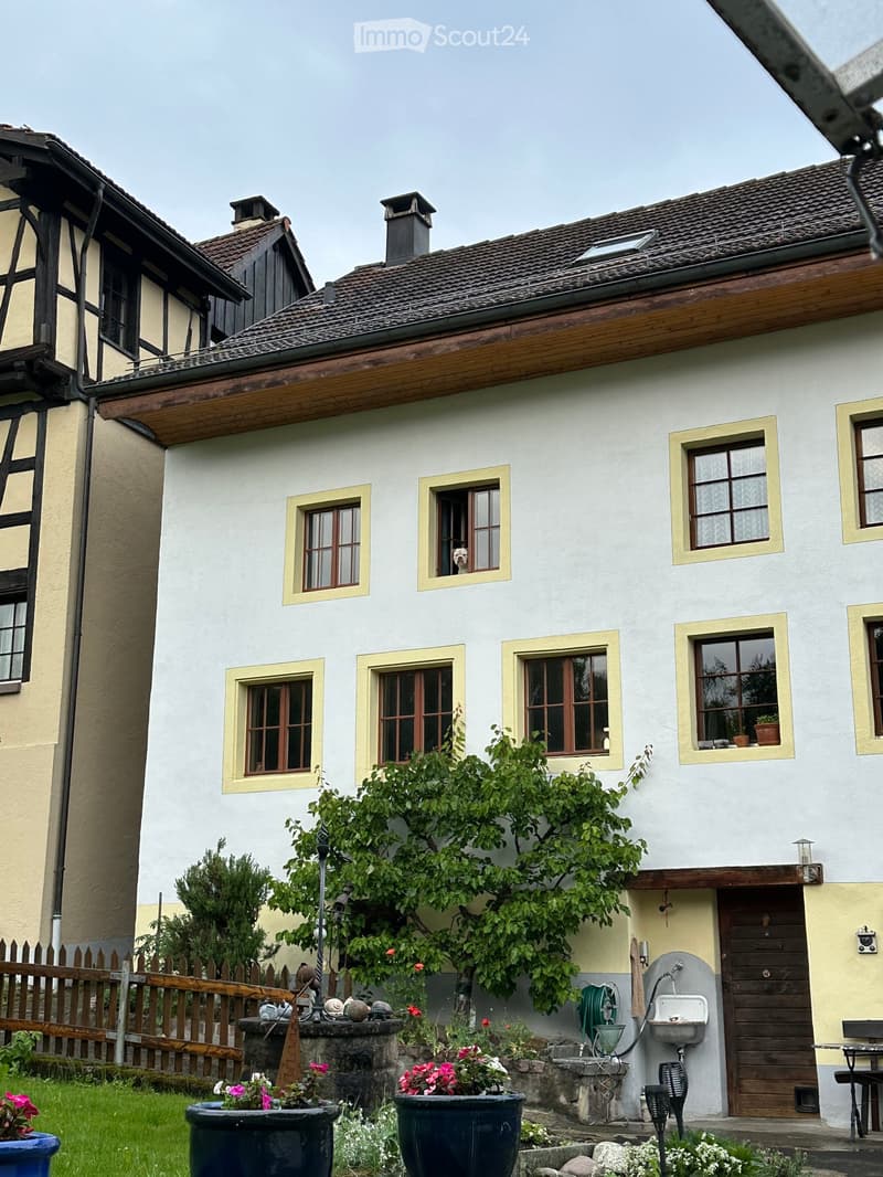 Two-story house with white walls, wooden trim, multiple windows, and a small garden area with potted plants and a wooden fence.