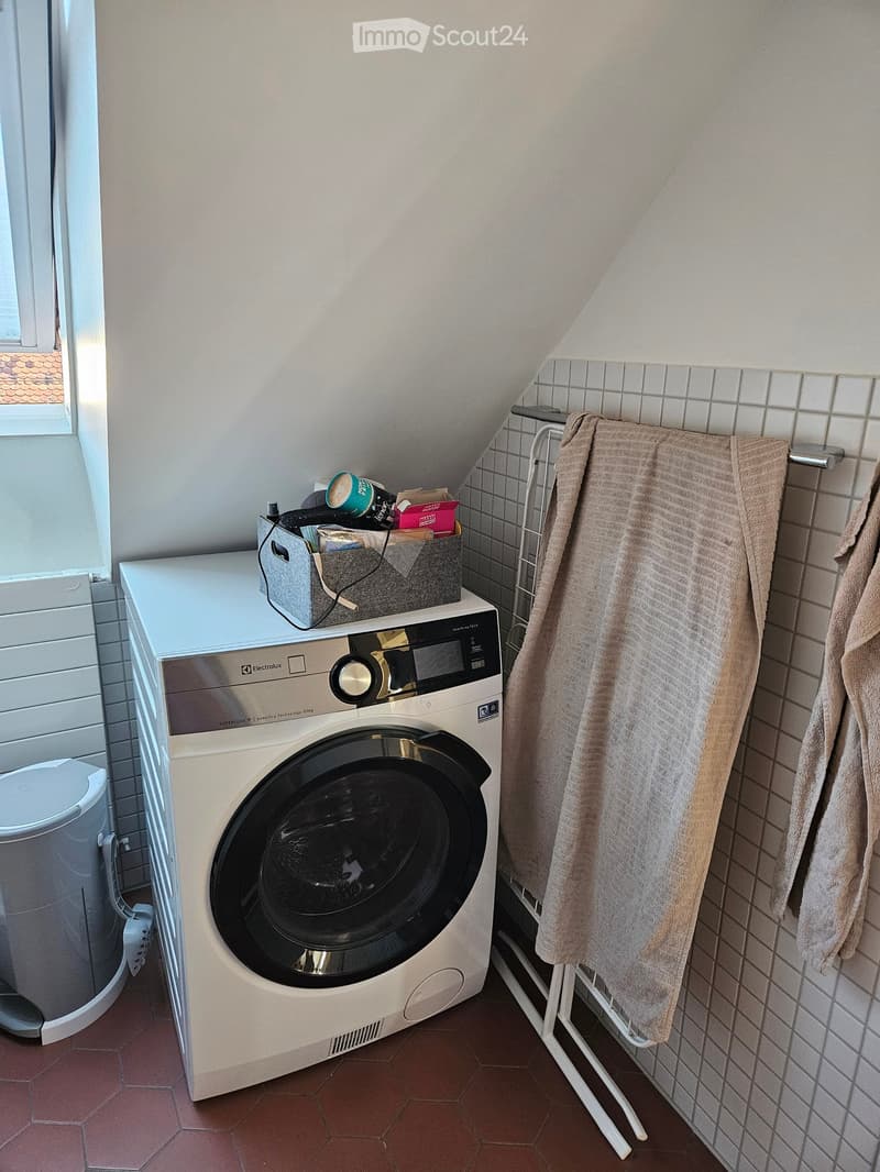 A small bathroom with a washing machine, a towel rack with towels, and a laundry basket on top of the washing machine.