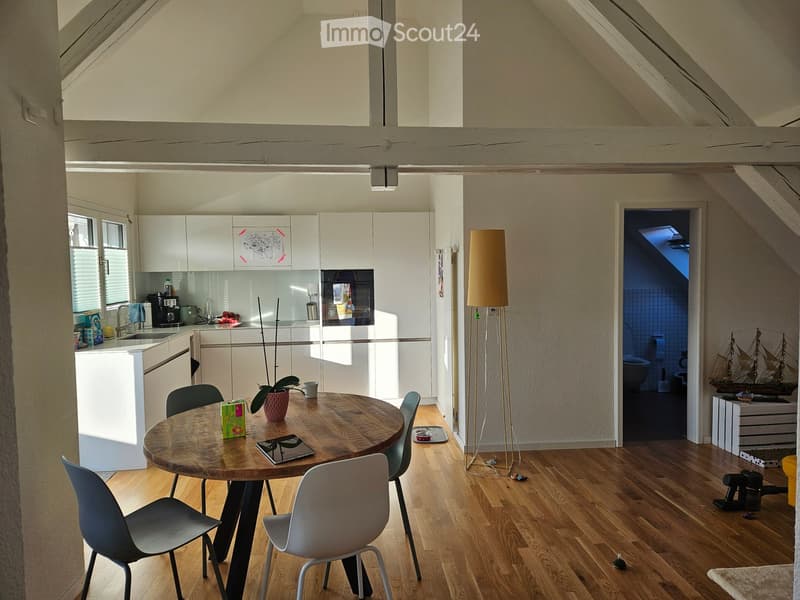 Bright, open-plan attic space with wooden floors, white cabinets, a round wooden dining table with chairs, a floor lamp, and a small kitchen area with a sink and refrigerator.