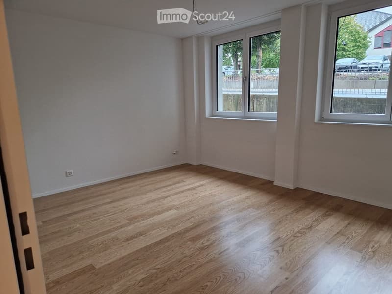 Empty room with wooden flooring and large windows, showing natural light and a view of the outside area.