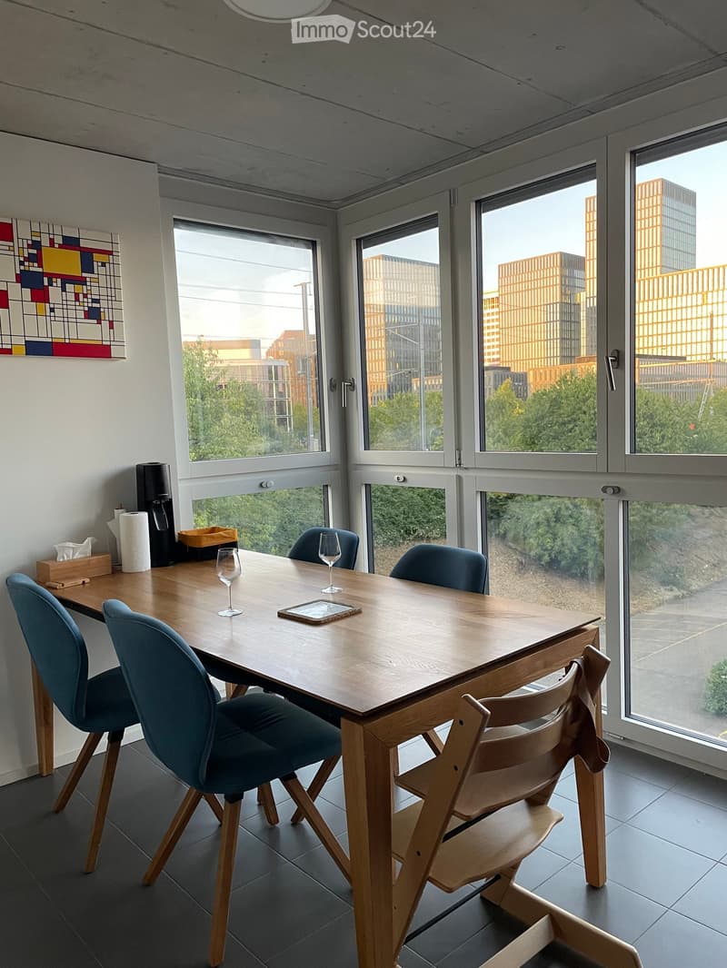 A modern, well-lit room with large windows overlooking a cityscape. The room features a wooden table surrounded by blue upholstered chairs and a wooden folding chair. On the table, there are two wine glasses and a tray. A small wooden cabinet holds paper