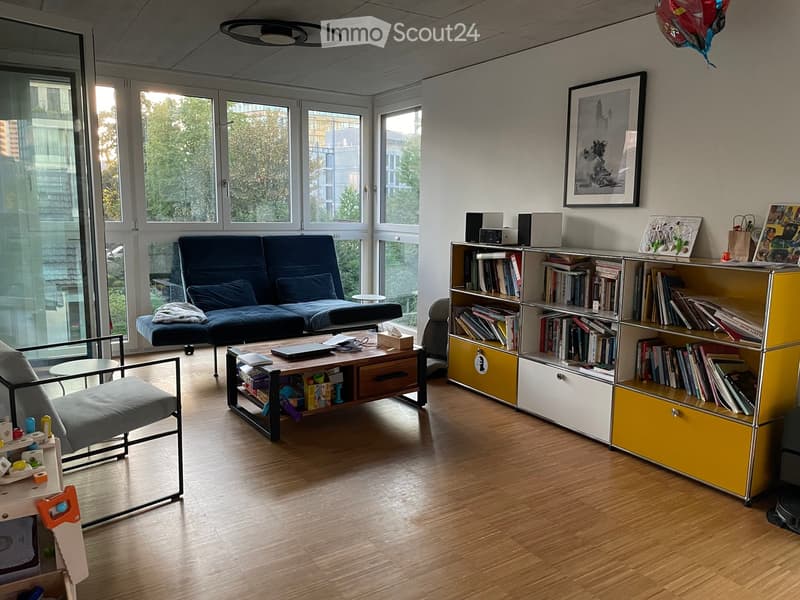 Modern living room with wooden flooring, large windows, a blue sofa, a coffee table, a bookshelf, and a chair.