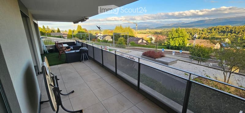 Balcony with tiled floor, metal railing, and scenic view of a town and mountains. Includes a wooden chair, table, and grill.
