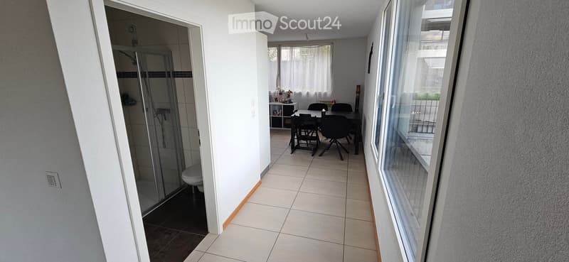 A modern, well-lit corridor leading to a dining area and a bathroom. The dining area features a black dining table with six black chairs, a small cabinet with a flower vase, and a window with white curtains. The bathroom includes a glass-enclosed shower a