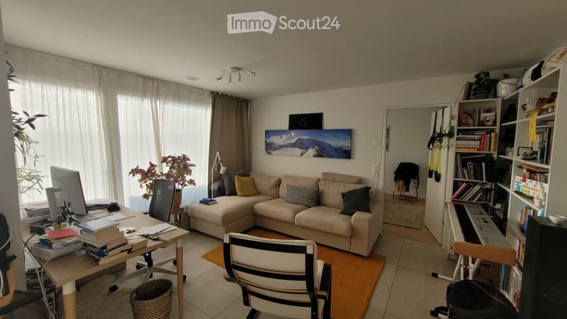 A well-lit living room with a beige sectional sofa, a wooden desk with a computer setup, a bookshelf filled with books, and a small piano. The room features large windows with curtains, a potted plant, and a colorful painting on the wall.