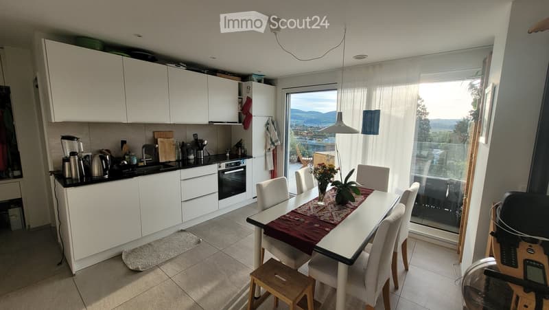 Modern kitchen with white cabinets, black countertops, and a dining area with a table and chairs. The room features a large window with a scenic view, a hanging pendant light, and various kitchen appliances.
