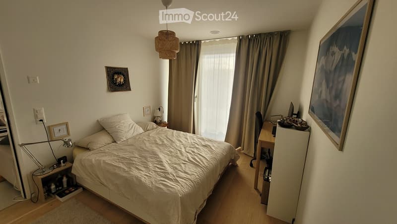 A modern bedroom with a large bed, nightstand, desk, and artwork on the walls. The room features wooden flooring, large windows with curtains, and a hanging pendant light.