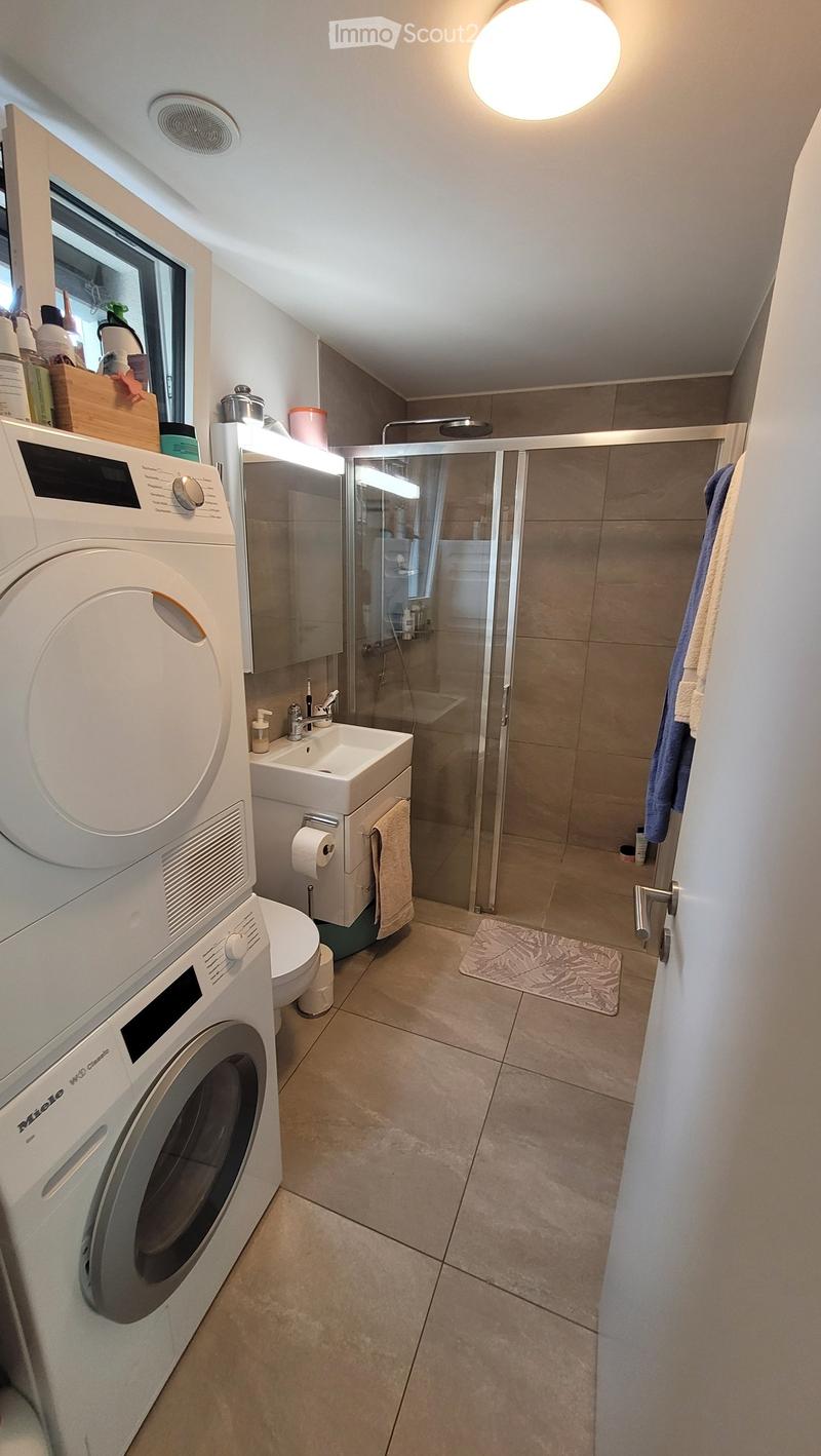 Modern bathroom with a washing machine, toilet, sink, and glass-enclosed shower. Features a towel, toilet paper, and a small bathroom mat.