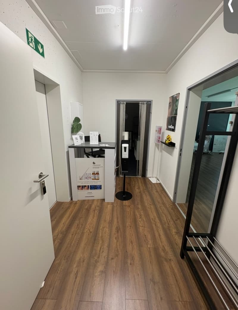 A clean, well-lit hallway with wooden flooring, white walls, and a reception area visible on the left side. There is a door with a silver handle, a small table with brochures, and a poster on the wall. A glass door with a metal frame is visible on the rig