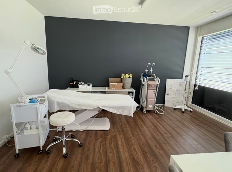 A well-lit, modern treatment room with a wooden floor, a treatment table, a white chair, a cabinet, a lamp, a vase with flowers, and a cosmetic device.