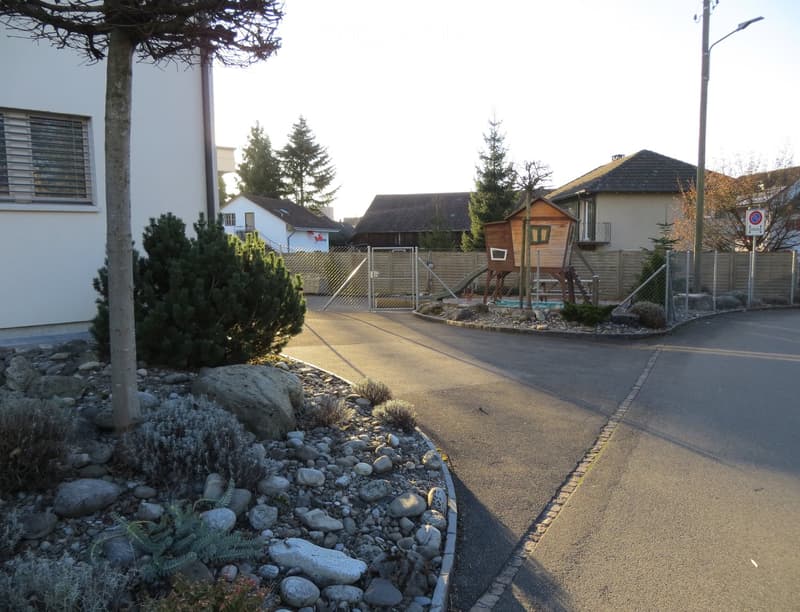 Residential driveway with landscaped garden area featuring rocks, shrubs, and small trees. A wooden playhouse and swing set are visible in the background, along with neighboring houses and a street sign.