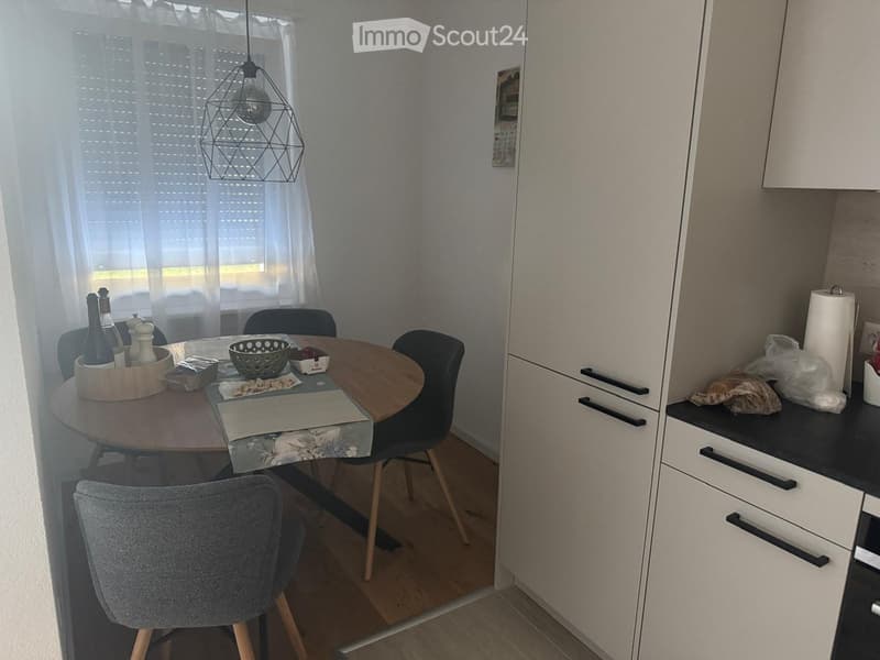 A modern kitchen with a round wooden dining table, four chairs, and a hanging geometric light fixture. The kitchen features white cabinets, a black countertop, and a window with white blinds.