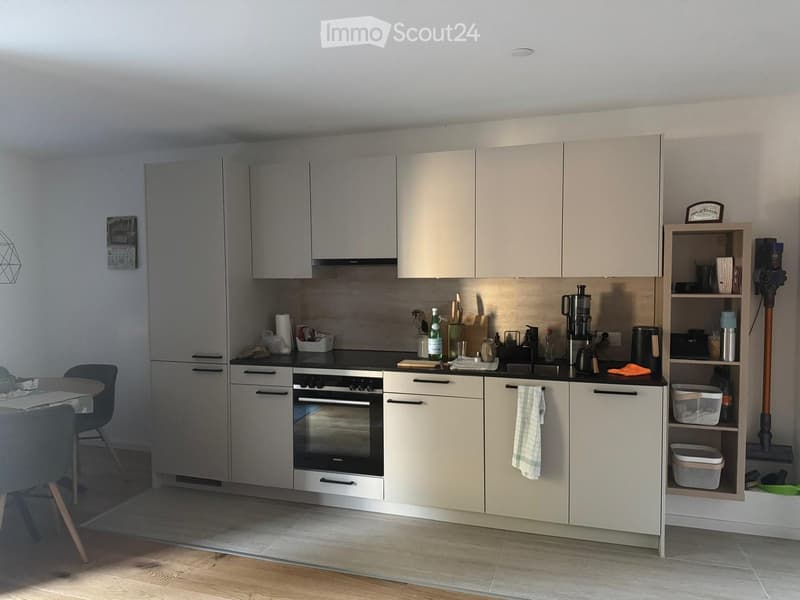 Modern kitchen with light-colored cabinets, stainless steel appliances, and a black countertop. Includes a refrigerator, oven, dishwasher, coffee maker, and a small dining area with a table and chairs.