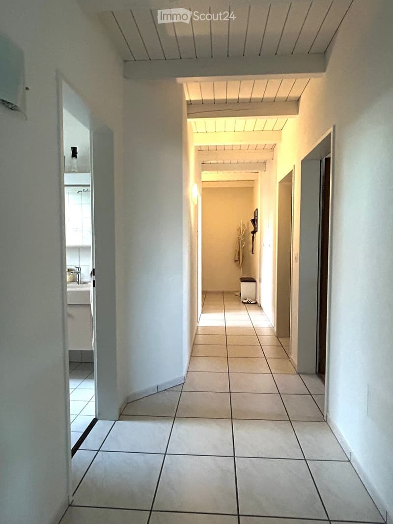 Clean, tiled hallway with white walls and a ceiling light fixture. Doors lead to adjacent rooms, including a bathroom and another hallway.