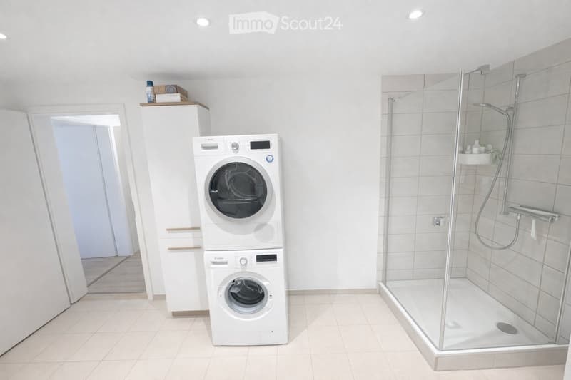 Modern bathroom with a white tiled shower stall, a stacked washing machine and dryer unit, and a small cabinet with a bottle on top.
