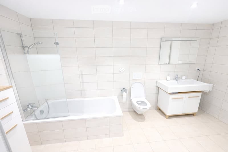 Modern bathroom with white tiles, a bathtub, toilet, sink, and a large mirror above the sink.