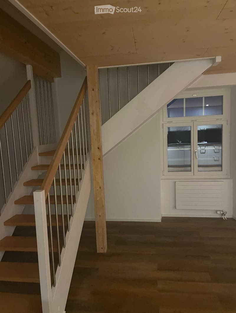 Empty hallway with wooden stairs, metal railing, and a window with glass panels. The floor is dark, and the walls are light-colored.