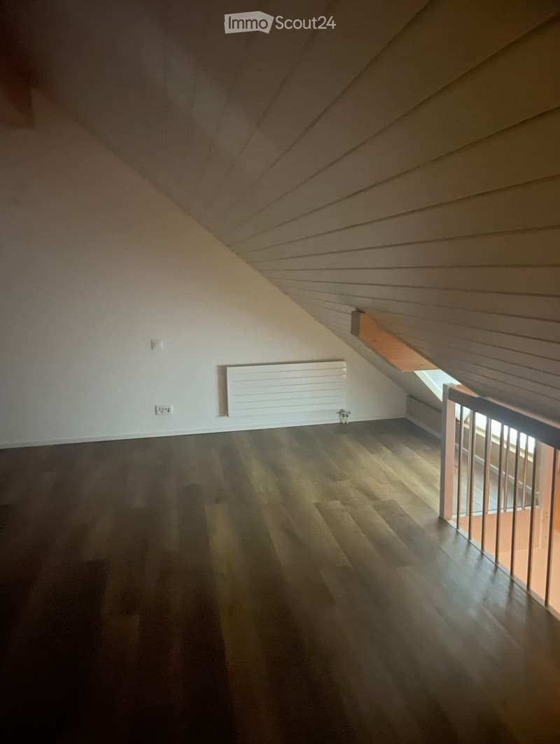 An empty attic space with wooden flooring, white walls, and a slanted wooden ceiling. A small window allows natural light to enter, and a white radiator is mounted on the wall.