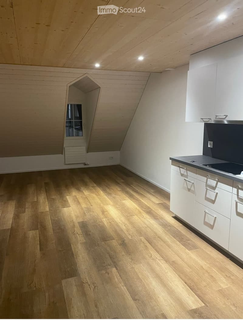 An unfurnished attic room with wooden flooring, white walls, and a small window. The room features built-in white cabinets and a countertop on the right side.