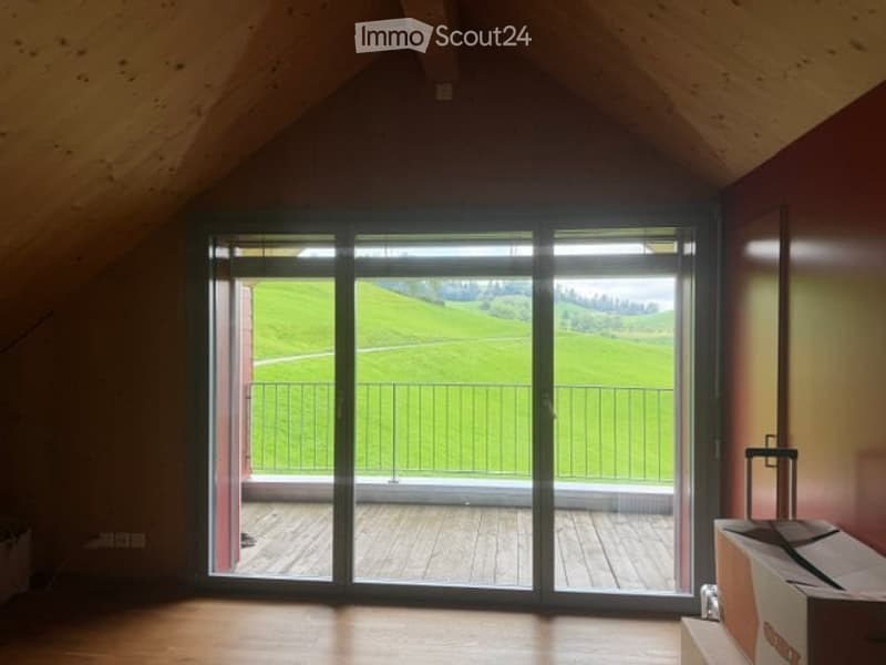 Empty attic room with wooden floors, large sliding glass doors opening to a wooden deck and scenic green landscape view.
