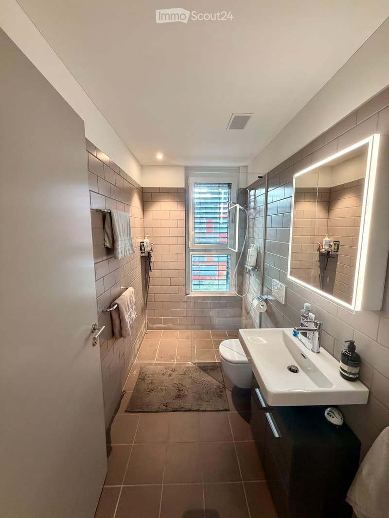 Modern bathroom with beige tiles, a large sink with a modern faucet, a toilet, a shower area with a glass enclosure, a large illuminated mirror, and towels on the wall.