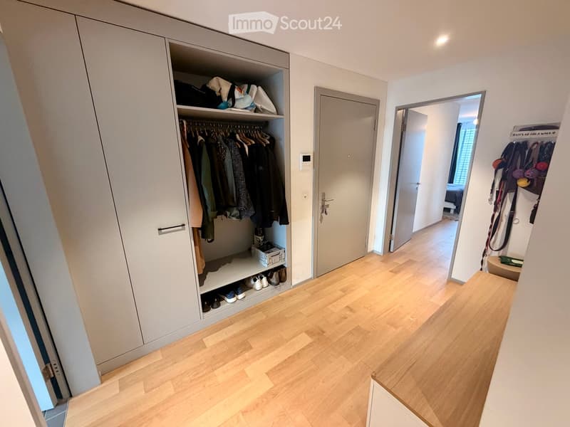 A modern hallway with light wooden flooring, white walls, and built-in closet with hanging clothes and shoes. A door leads to another room, and a small shelf with items is visible in the background.