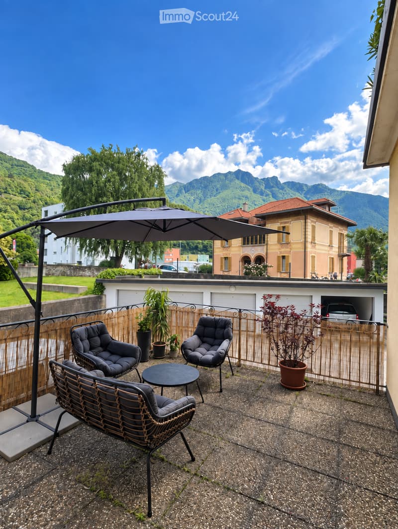 Balcony with a view of mountains, featuring a black umbrella, three chairs, a small table, and potted plants.
