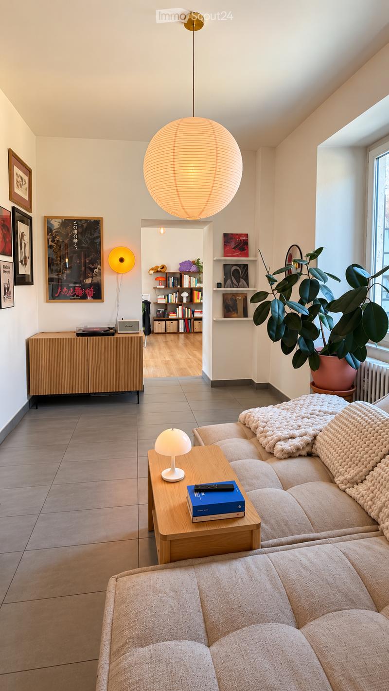 Modern living room with a light-colored sofa, a wooden coffee table, a small table lamp, a bookshelf, and a potted plant near a window. The room features a large spherical pendant light and several picture frames on the walls.