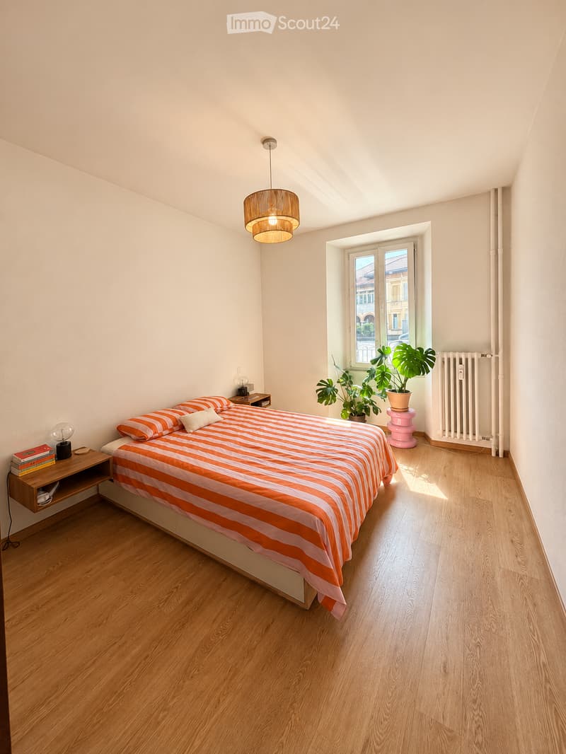 Bright bedroom with wooden flooring, a large bed with orange and white striped bedding, a nightstand with a table lamp and books, a potted plant near the window, and a wicker pendant light hanging from the ceiling.