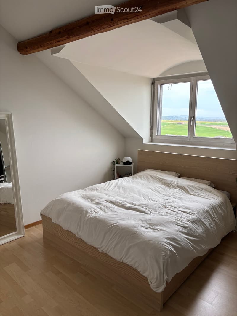 Bright attic bedroom with wooden bed, white bedding, nightstand, and large window overlooking a green field.
