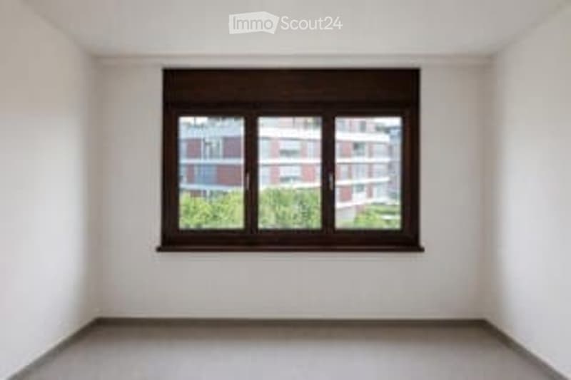 Completely empty room with white walls, grey floor, and a single window with black frame and blinds.