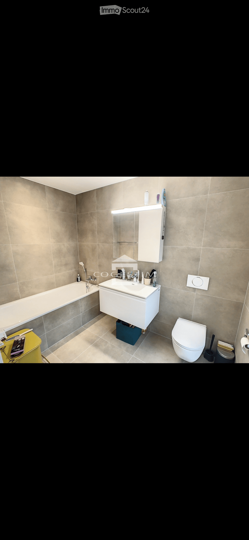 Modern bathroom with beige tiled walls and floor, featuring a bathtub, sink with mirror cabinet, toilet, and various toiletries.