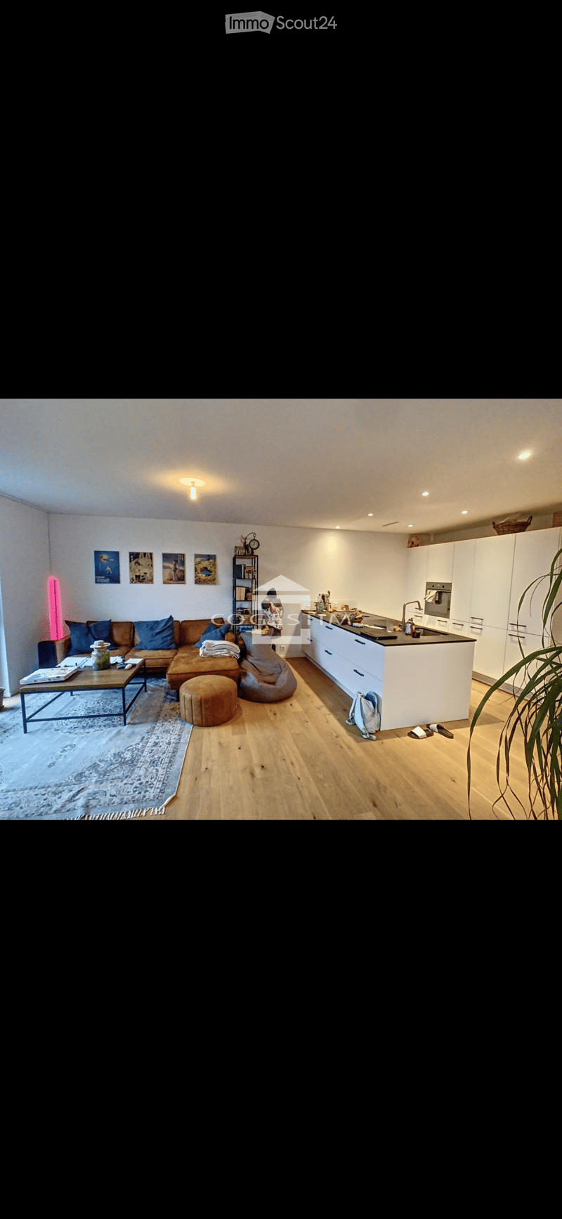 Open-plan living and dining area with a modern kitchen, featuring a brown sectional sofa, coffee table, beanie chair, and a kitchen island with white cabinets and a black countertop.