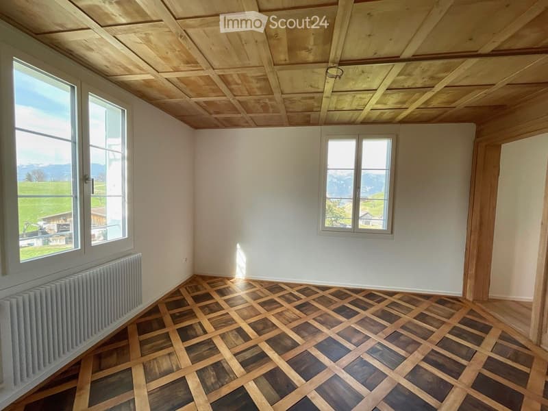 Empty room with wooden parquet flooring, white walls, exposed wooden ceiling beams, and large windows offering a scenic mountain view.