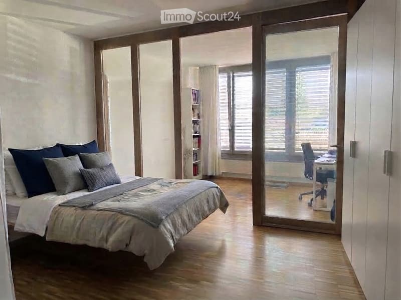 Modern bedroom with a large bed, multiple pillows, and a grey duvet. A sliding glass door leads to a balcony area with a desk and chair. Wooden flooring and white walls.