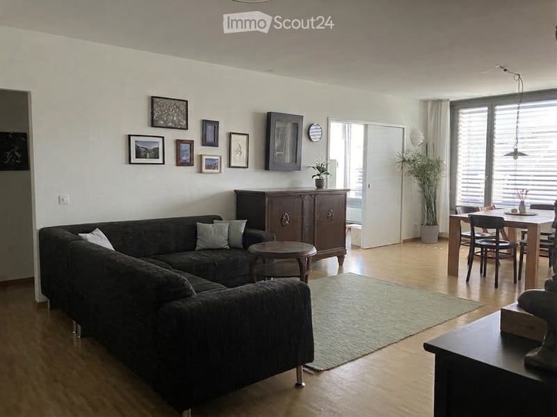 Modern living room with a dark grey sectional sofa, wooden coffee table, dining area with a table and chairs, and a large window with blinds. The room features a light-colored wooden floor, a green area rug, and several picture frames on the wall.