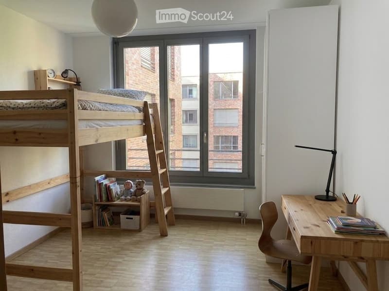 Bright, modern bedroom with a wooden bunk bed, desk, chair, and small shelf holding books and toys. Large window offers a view of the building outside.
